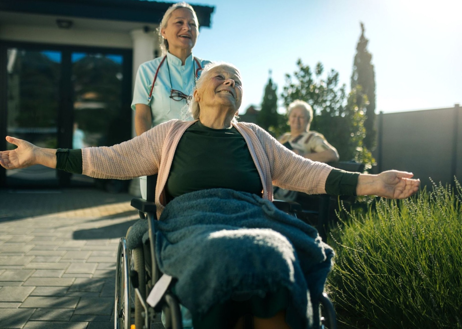 Eine Begleiterin schiebt eine ältere Frau im Rollstuhl durch einen sonnigen Garten, die Frau breitet die Arme aus